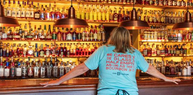 A woman in a light blue shirt stands in front of seemingly hundrands of bourbon botttles. Her shirt reads, "Who cares if the glass is half empty as long as there's bourbon in it" The B-Line