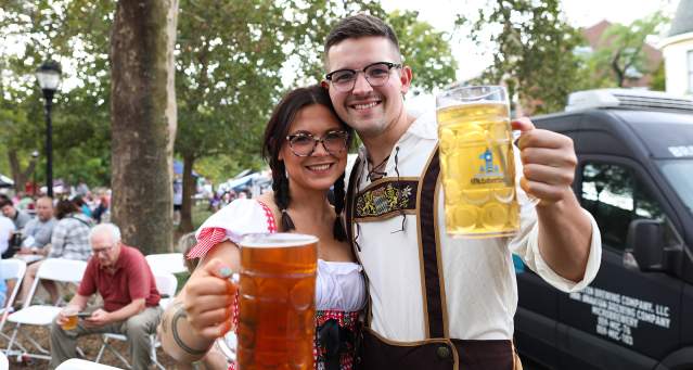 A smiling couple hoist beer steins to the camera.