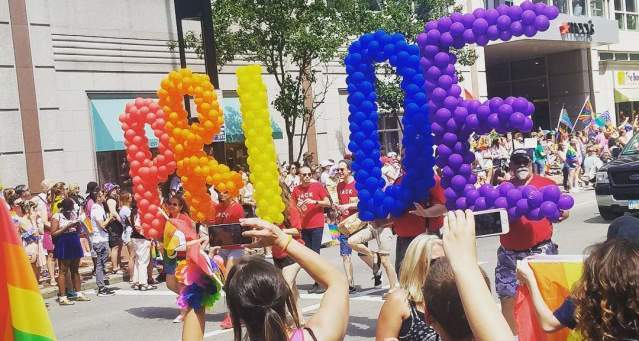 Image is of the Cincinnati Pride Parade with a group carrying the letters P.R.I.D.E made of ballons as they walk down the street.
