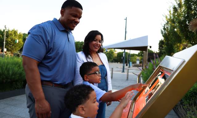 A family interacts with an outdoor display at the Black Music Walk of Fame in the foreground, while a park with trees and a covered structure is visible in the background.