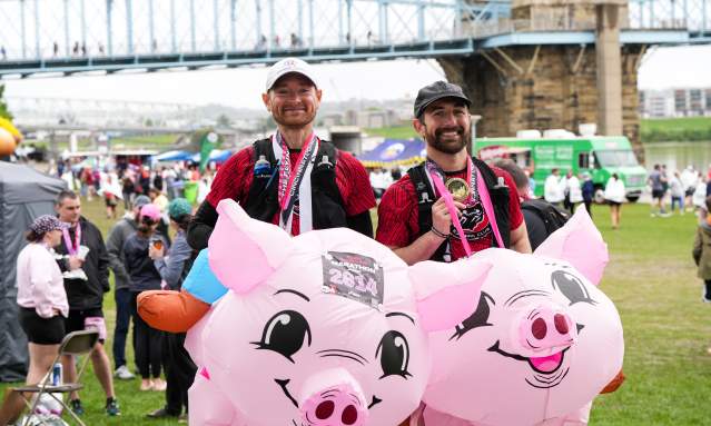 Two men after Flying Pig with medals in flying pig costumes in front of the Roebling Bridge