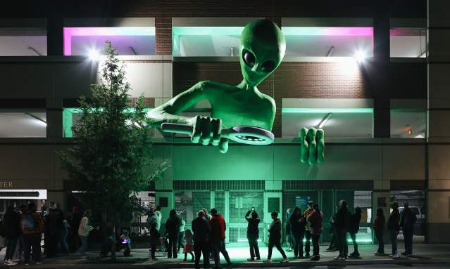 A large alien sculpture looks over the crowd on the sidewalk under it.