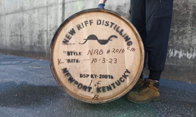 A New Riff barrel sits on its side with the distillery stamp visible. A person stands next to the barrel to steady it.