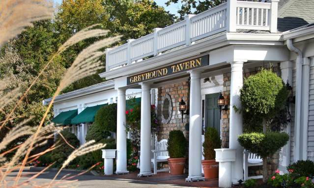 Exterior of Greyhound Tavern entrance. White columns flank the porch dotted with green bushes.