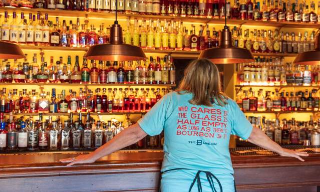 A woman in a light blue shirt stands in front of seemingly hundrands of bourbon botttles. Her shirt reads, "Who cares if the glass is half empty as long as there's bourbon in it" The B-Line