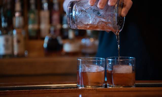 A close up of two drinks being poured over large, square ice in rocks glasses.