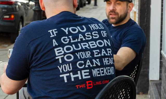 A man sits facing away from the camera. The back of his shirt reads "If you hold a glass of bourbon to your ear you can hear the weekend". Behind him is another man, facing the camera and looking at the first man.