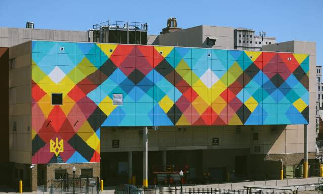 A large-scale mural on the side of a convention center features colorful criss-crossing line reminiscent of a weaving