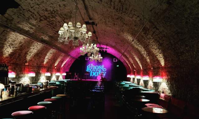 A tunnel lit with white and pink lights with a sign at the end in blue reading ghost baby. Rows of barstools line the walls of the tunnel