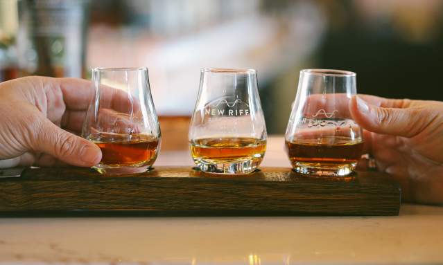 Three glencairn glasses, each with a pour of whiskey, sit in a wooden holder. Hands from either side reach out to select a glass.