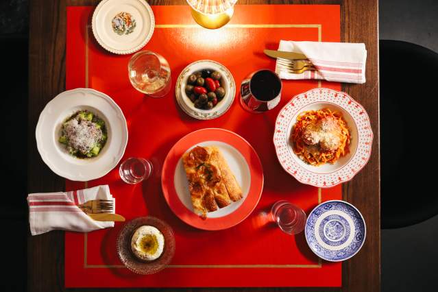 A red table is set with two place settings. One has a pasta with red sauce, the other a pasta with pesto and cheese. A bowl of olives and a plate of bread are between the two settings.