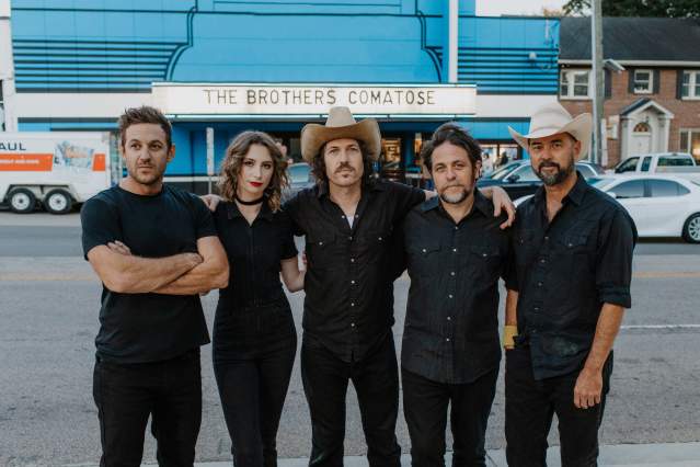 A group of people dressed in black face the camera. Behind them a blue building has a marquee that reads "The Brothers Comatose".