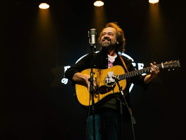 A man stands in front of a microphone, holding an acoustic guitar.