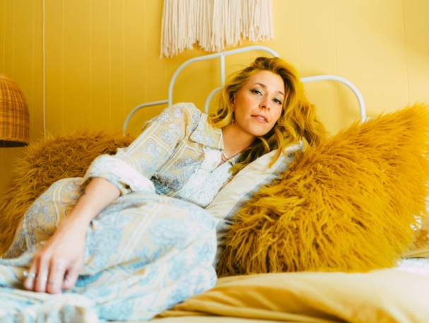 A women in a light blue and white outfit lays on a bed, leaning away from the camera. The bedding, the walls, and her hair are various shades of yellow.