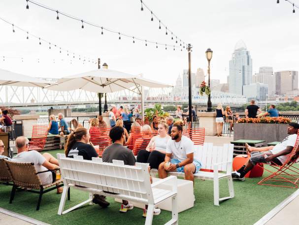 A group of people enjoy outside at Bridgeview Box Park. The Cincy skyline is in the background.