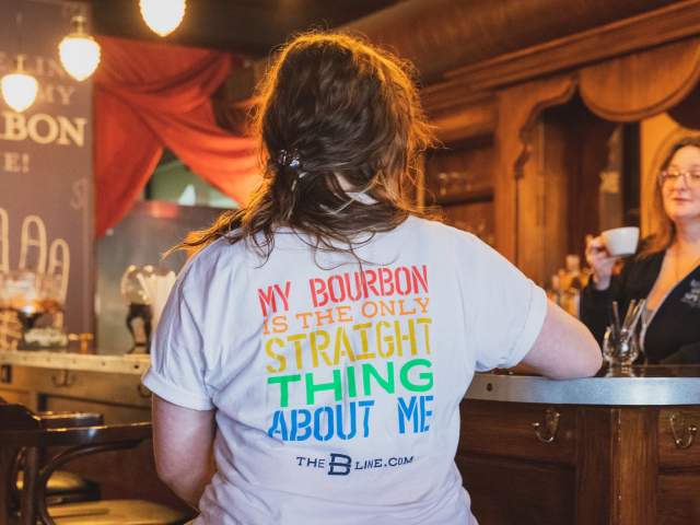 A person sits facing away from the camera in a white shirt. In rainbow order the shirt says "My Bourbon is the only straight thing about me"
