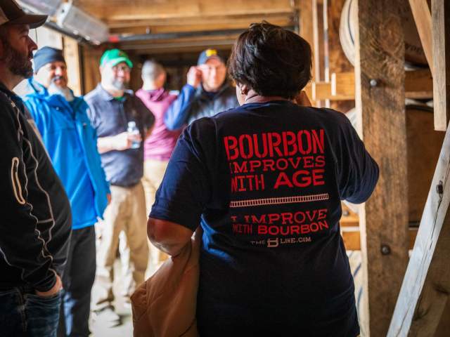 A woman faces away from the camera, speaking to a tour group. Her shirt says "Bourbon improves with age, but I improve with bourbon".