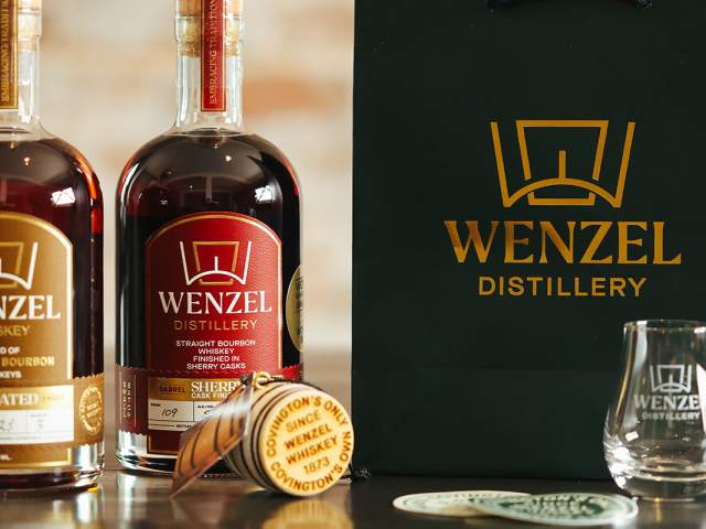Two bourbon bottles sit on a table next to a bag with the Distillery logo on it. A small wooden barrel says "Covington's Only, Covington's Own, Since1873, Wenzel Whiskey"