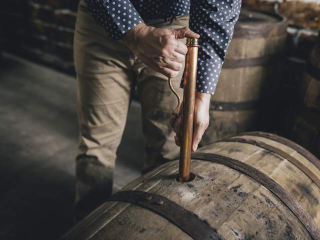 A person in khaki pants and a blue shirt uses a long copper tube, called a whisky thief, to remove bourbon from a barrel laying on its side.