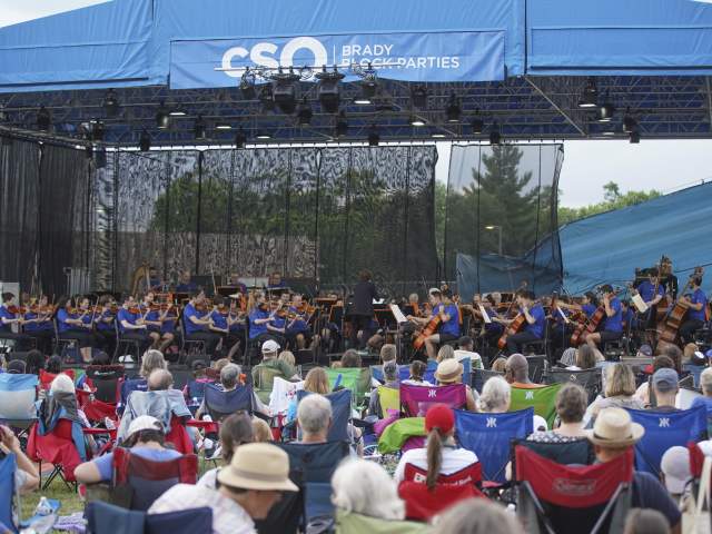A crowd of people in pop-up lawn chairs sit by a stage and watch the Cincinnati Symphony Orchestra perform