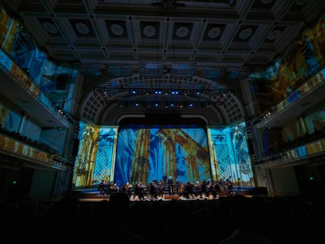 The Cincinnati Symphony Orchestra plays in front of a lit up back drop.