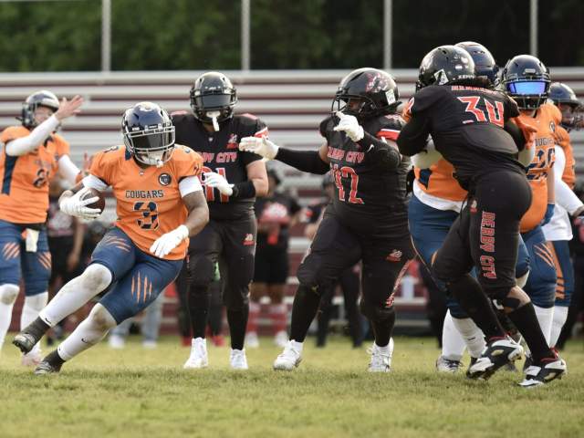 A player in orange and blue cradles a football while running past players in black.
