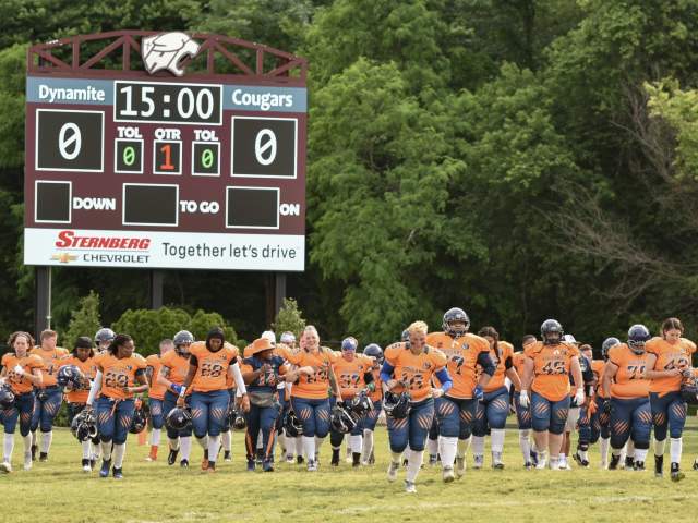 The football team in orange and blue uniforms enter the field at the start of a game. The scoreboard is in the background.