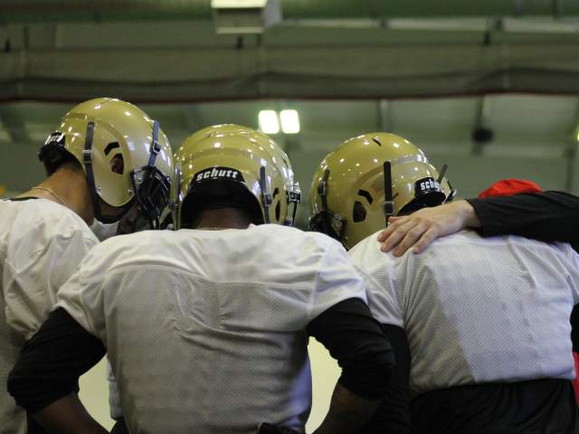 Three players huddle together with their backs to the camera. They are wearing white jerseys and gold helmets.