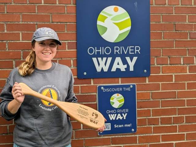An Ohio River Way representative holds a paddle in front of an official designation/trail sign