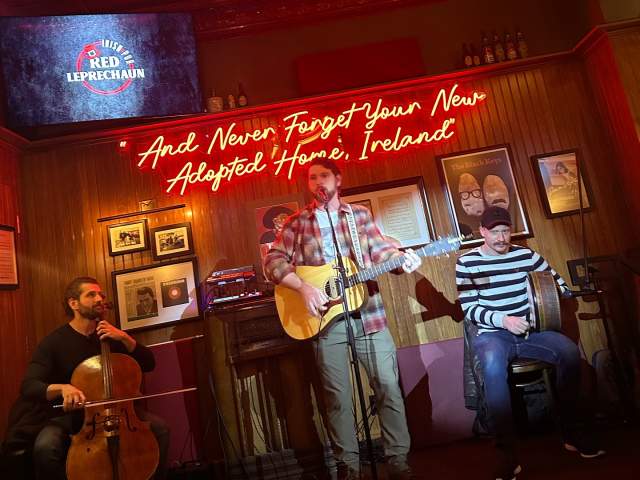 A music group plays on a stage. A neon sign above them says "And never forget your new adopted home, Ireland".
