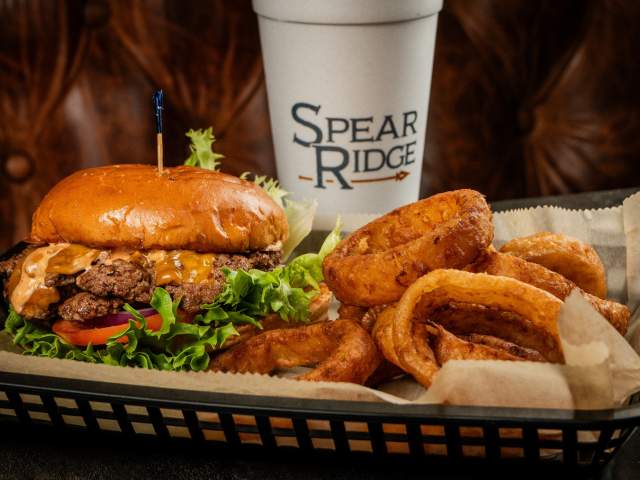 A double cheeseburger sits in a basket next to an order of golden onion rings. The cup in the background says "Spear Ridge".