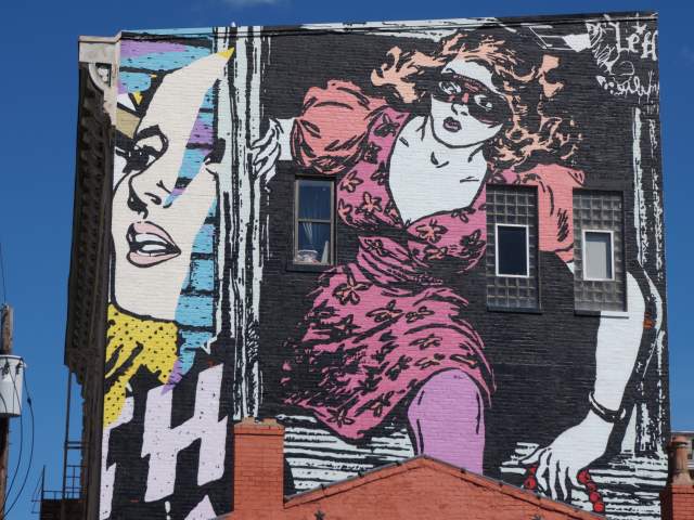 A street art mural by Faile in Covington, Ky. of a woman in a pink dress, wearing a black mask, climbing out of a building