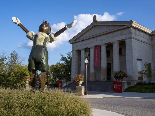 Image is of the Cincinnati Art Museum entrance in the background with a bronze statue of Pinocchio with his hands raised looking to the sky.