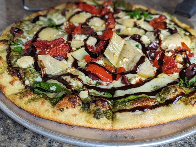 The image is of a Mediterranean pizza featuring a pesto sauce, blend of Italian cheeses, tomatoes and a balsamic glaze.