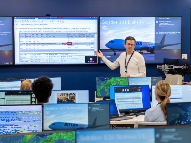 People sit at computers listening to a person speak in the front of the room. There are multiple screens.