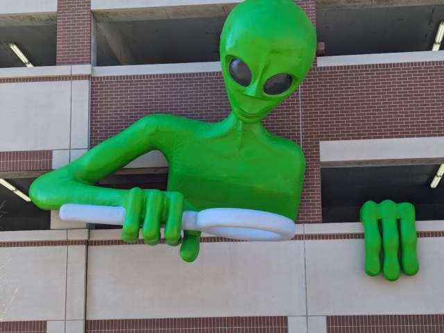 A sculpture of a 30 foot green alien leaning out of a parking garage and peering through a magnifying glass