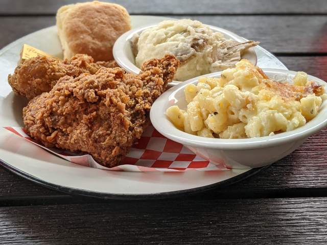 Crispy fried chicken, mashed potatoes, and macaroni and cheese are served on a plate with a biscuit, all set against a dark wooden table.