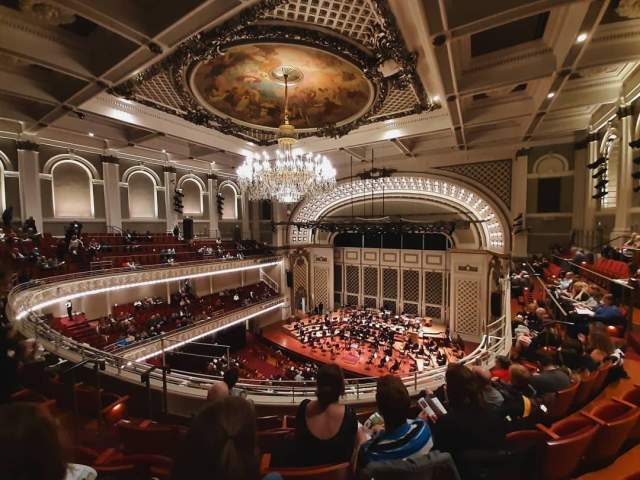 Inside an amphitheater from the balcony. White lights frame the stage and line the edges of the balconies. A small orchestra is playing to an audience