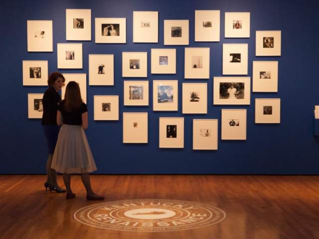 Two people stand in front of a dark blue wall. Hung in a cluster are many framed photographs with large mats.
