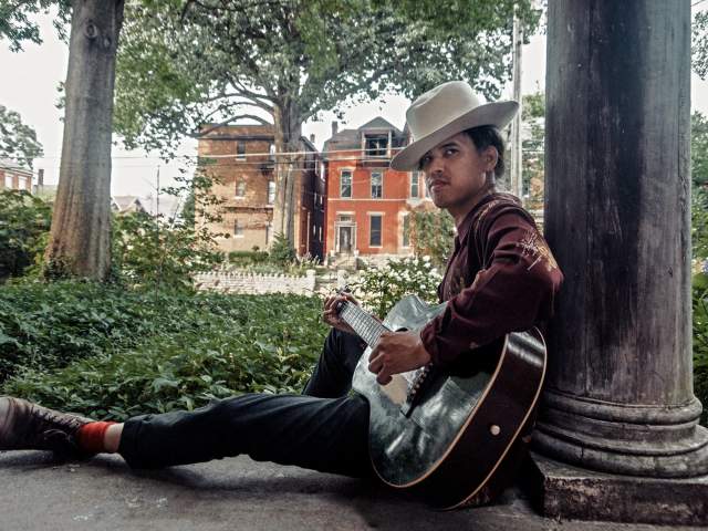 A man in a cowboy hat looks past the camera. He's holding a guitar and is leaning against a pillar.