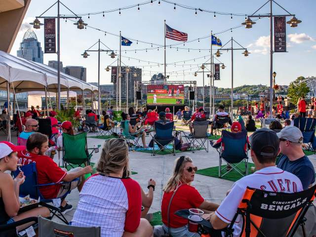A crowd of people in Cincinnati Reds clothing watching the Opening Day Reds game on a big screen at Newport on the Levee