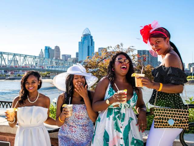 Four women in festive Derby attire enjoy drinks by the Ohio River with the Cincinnati skyline in the background.