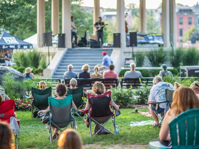 An audience sits on chairs and blankets watching a live performance at the Gazebo in Washington Park.