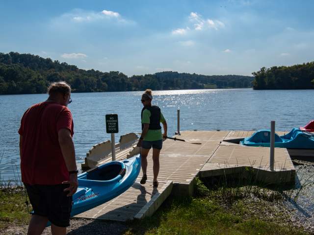 Two people carry a kayak on to a small dock with a kayak launch.