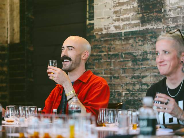 A male and female sitting against a brink wall tasting bourbon from a barrel pick.