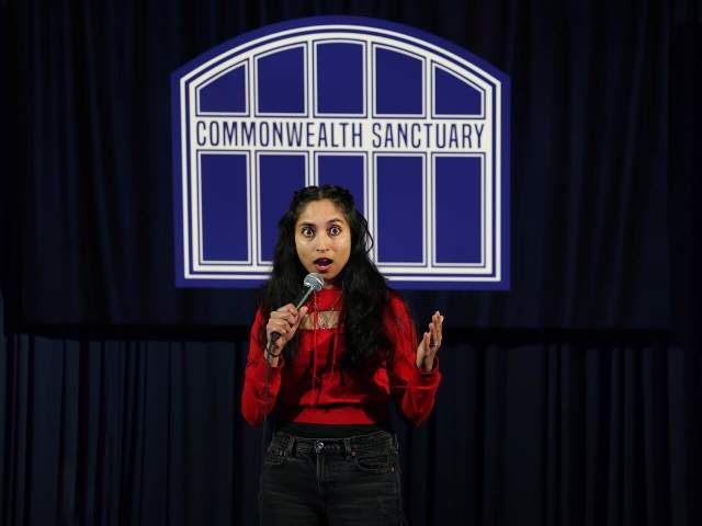 A woman in a red shirt holds a microphone on stage in front of a sign that says 'Commonwealth Sanctuary'.