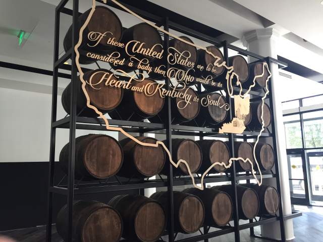 A rack of bourbon barrels with a gold, stenciled map of the U.S.and the words "If the United States are to be considered a body then Ohio would be its heart and Kentucky its soul.