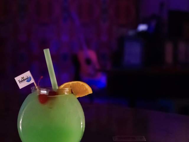 A green drink served in a fishbowl shaped glass, garnished with an orange slice and cherry.