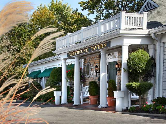 Exterior of Greyhound Tavern entrance. White columns flank the porch dotted with green bushes.