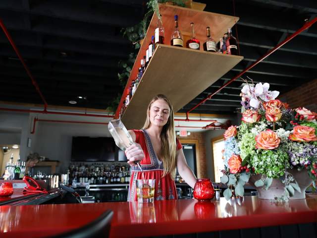 A woman in a red dress makes a drink at the red, wood bar next to a vase of flowers. A shelf of bottles is suspended above her.